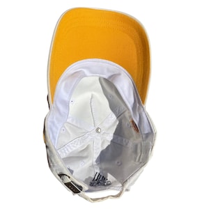 May include: A white baseball cap with a yellow brim. The underside of the cap is white, with the Adidas logo visible. The cap has a metal clasp for adjustment.