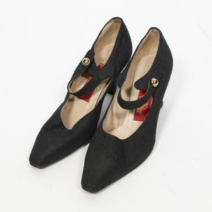 May include: A pair of black fabric Mary Jane style shoes with pointed toes and small heels. Each shoe has a strap across the top with a gold-colored button closure. The interior of the shoe is a light beige color.