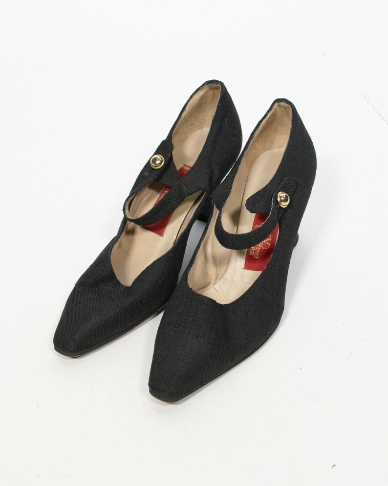 May include: A pair of black fabric Mary Jane style shoes with pointed toes and small heels. Each shoe has a strap across the top with a gold-colored button closure. The interior of the shoe is a light beige color.