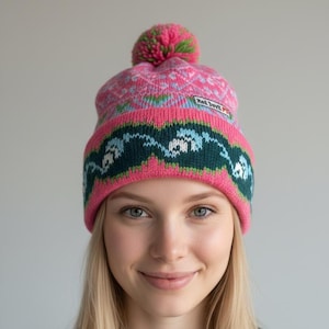 Puede incluir: Un gorro de punto rosa con un pompón verde y rosa. El gorro presenta un diseño estampado en rosa, turquesa y blanco, con una banda verde oscuro. El texto "Red Devil P" es visible en el gorro.