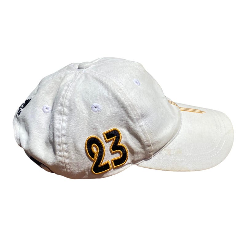 May include: White baseball cap with the number 23 in black and gold on the side. The cap has a curved brim and the Adidas logo on the side. The cap is made of a soft, durable material.