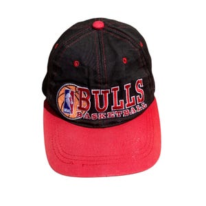 Può includere: Cappellino da baseball nero e rosso con il logo dei Chicago Bulls. Il cappellino ha una visiera rossa e una corona nera con cuciture rosse. Il logo dei Bulls è ricamato sul davanti, con il logo NBA sul lato sinistro del pallone da basket.