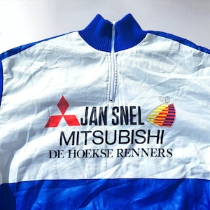 Puede incluir: Un maillot de ciclismo vintage blanco y azul con cierre de media cremallera. El maillot presenta el texto "JAN SNEL MITSUBISHI DE HOEKSE RENNERS" en negro, un logotipo rojo y un emblema colorido. El cuello y las mangas son azules.