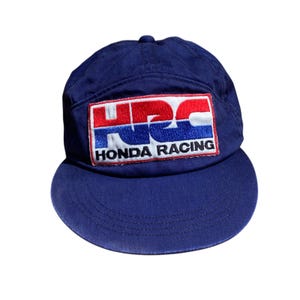 Könnte beinhalten: Marineblaue Baseballkappe mit gebogenem Schirm. Die Kappe hat einen rechteckigen Aufnäher mit dem Text "HRC HONDA RACING" in Weiß, Rot und Blau. Die Kappe ist aus strapazierfähigem Stoff gefertigt und hat ein klassisches, sportliches Design.