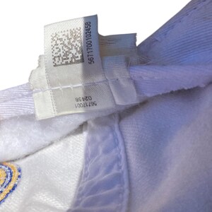May include: Close-up of a white baseball cap with a tag. The tag has the numbers "5671700102456" and "56717001 0245B" printed on it. The cap is made of a soft, textured material.