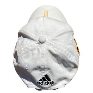 May include: White baseball cap with the Adidas logo in black on the front. The cap has a curved brim and a button on top. Gold vertical stripes are on the back of the cap.