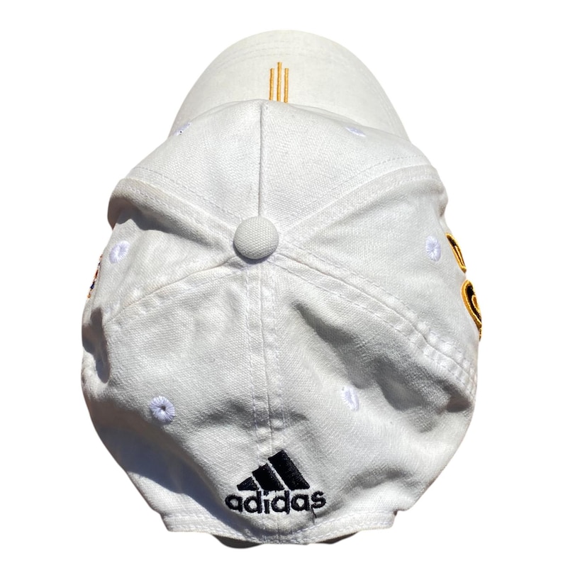 May include: White baseball cap with the Adidas logo in black on the front. The cap has a curved brim and a button on top. Gold vertical stripes are on the back of the cap.
