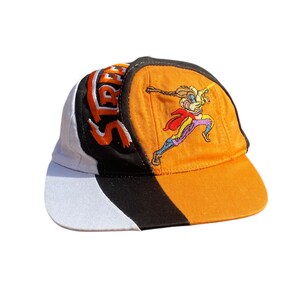 Puede incluir: Una gorra de béisbol multicolor con paneles blancos, negros y naranjas. La parte frontal muestra un gráfico bordado de un personaje corriendo con capa roja, con la palabra STREET bordada en letras naranjas en el panel lateral.