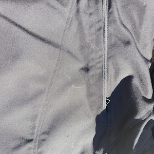 May include: Close-up of black fabric with visible stitching and a zipper. The fabric appears to be a durable material, possibly for outerwear. The image is well-lit, highlighting the texture and details of the garment.