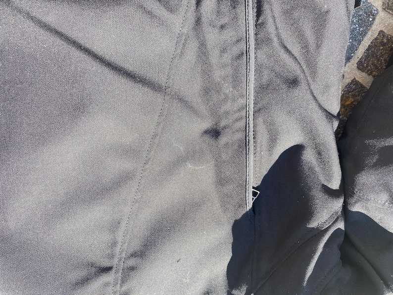 May include: Close-up of black fabric with visible stitching and a zipper. The fabric appears to be a durable material, possibly for outerwear. The image is well-lit, highlighting the texture and details of the garment.