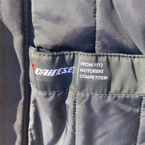 May include: Close-up of a black quilted fabric with a label. The label features the brand name "DAINESE" in white, with a red accent. Below the brand name, the label reads "FROM 1972 MOTORBIKE COMPETITION" in white text.