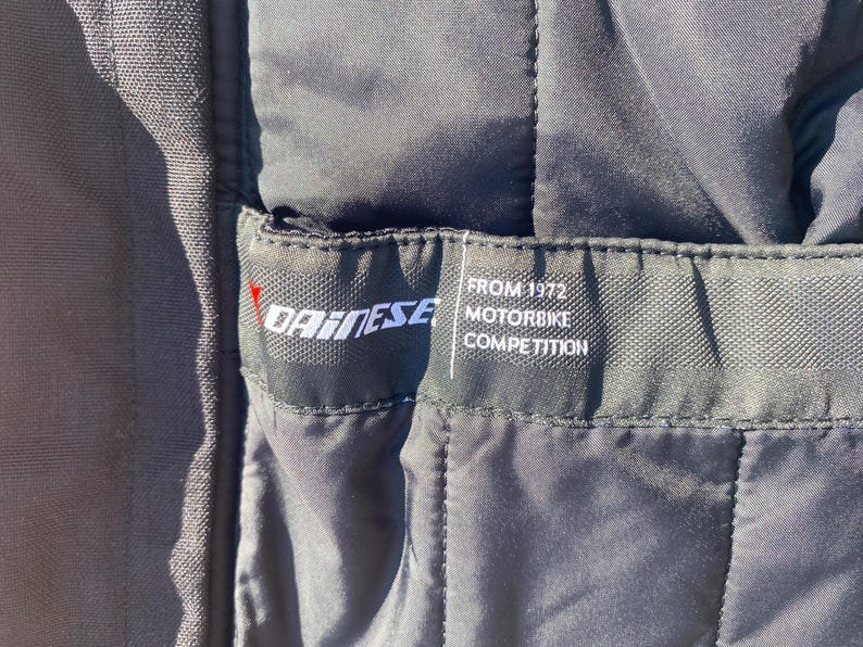 May include: Close-up of a black quilted fabric with a label. The label features the brand name "DAINESE" in white, with a red accent. Below the brand name, the label reads "FROM 1972 MOTORBIKE COMPETITION" in white text.