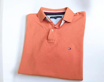 May include: An orange polo shirt with a collar and short sleeves. The shirt has a two-button placket and a small logo on the chest. The shirt is folded neatly on a white surface.