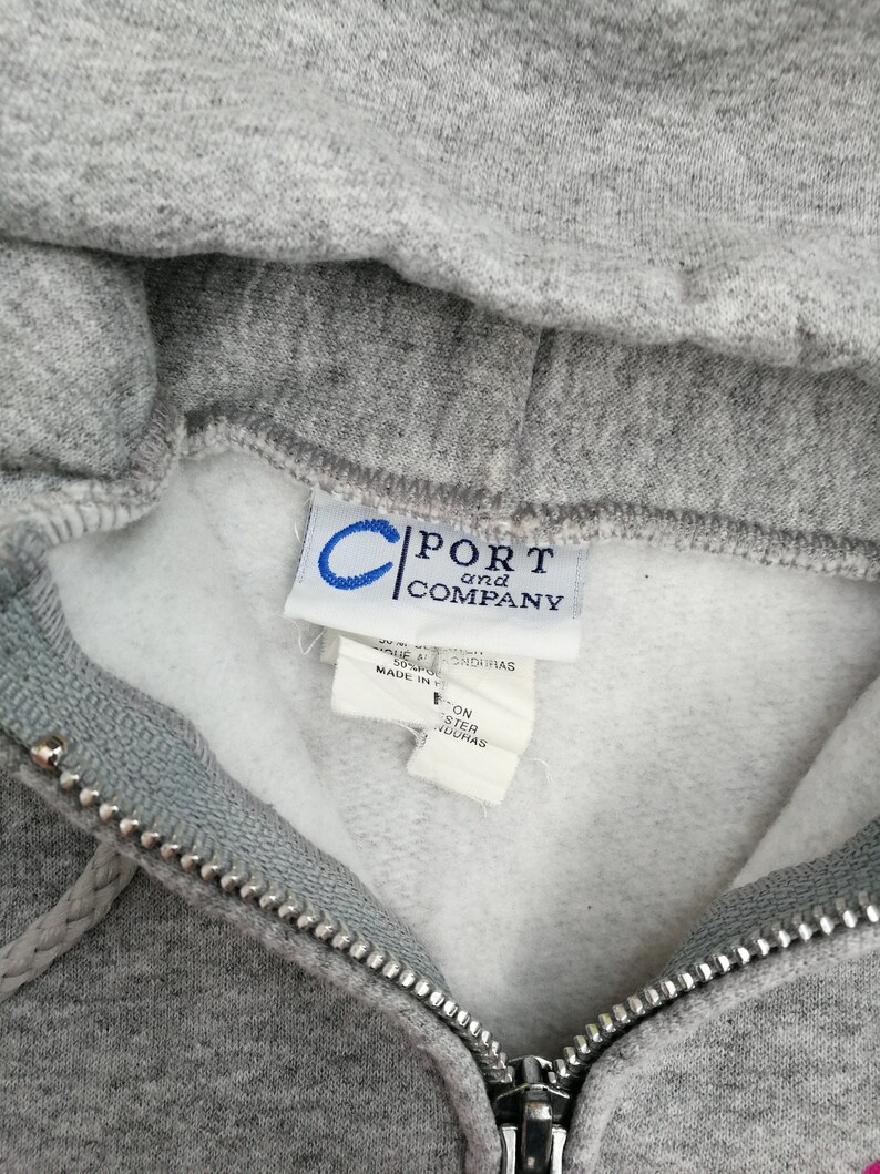 May include: Gray hooded sweatshirt with a zipper closure. The tag reads "Port and Company" and indicates the garment is made in Honduras.
