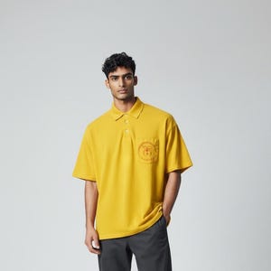May include: Mustard yellow polo shirt with a chest pocket featuring an embroidered design. The shirt has a classic collar and short sleeves. The model is wearing dark gray pants.