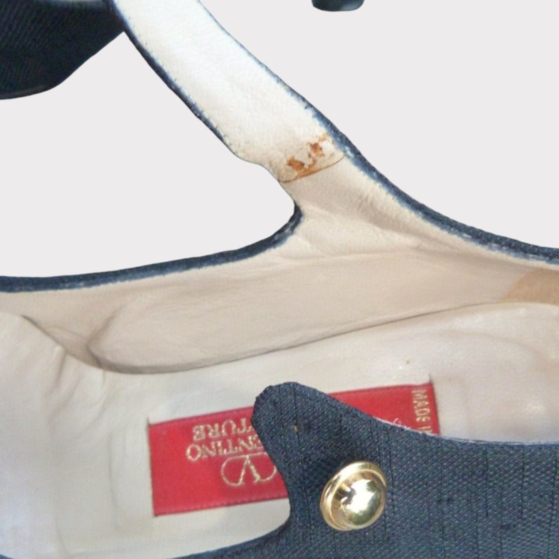May include: Close-up of a navy blue shoe interior with a white leather insole. A red tag with gold text is visible. The shoe has a gold button detail.