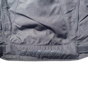 May include: Close-up of a black motorcycle jacket with the brand name "DAINESE" embroidered in a darker shade of black. The jacket has visible seams and a textured surface, suggesting a durable material.