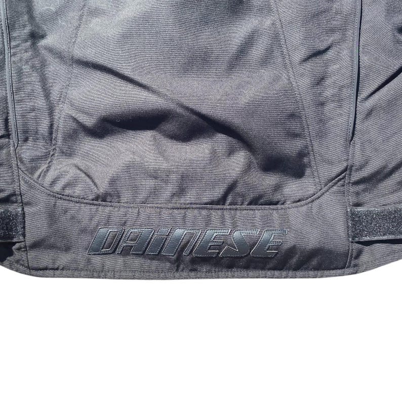May include: Close-up of a black motorcycle jacket with the brand name "DAINESE" embroidered in a darker shade of black. The jacket has visible seams and a textured surface, suggesting a durable material.
