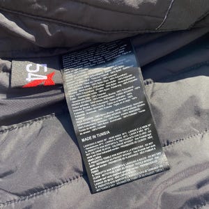 May include: Close-up of a black garment label with white text and a red logo. The label is attached to a dark gray quilted fabric. The label includes washing instructions and material composition.