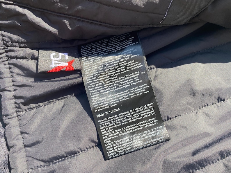 May include: Close-up of a black garment label with white text and a red logo. The label is attached to a dark gray quilted fabric. The label includes washing instructions and material composition.