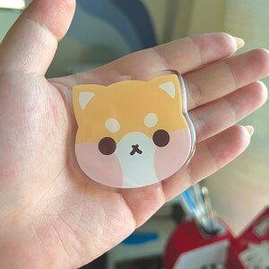 May include: A clear acrylic keychain featuring a cartoon Shiba Inu dog design. The dog is yellow with pink cheeks and brown eyes.