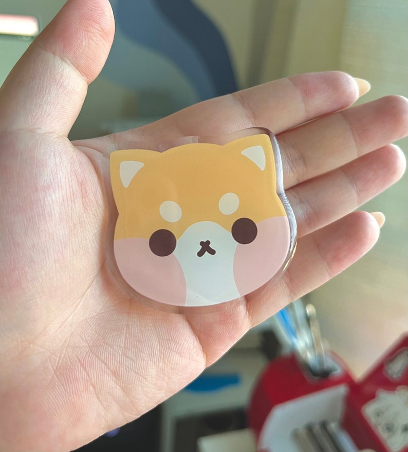 May include: A clear acrylic keychain featuring a cartoon Shiba Inu dog design. The dog is yellow with pink cheeks and brown eyes.