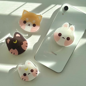 May include: Four phone grips with cute animal faces. The grips are clear plastic with a white background and pink cheeks. The animals are a brown and white dog, a black and white cat, a white cat, and a white bunny.