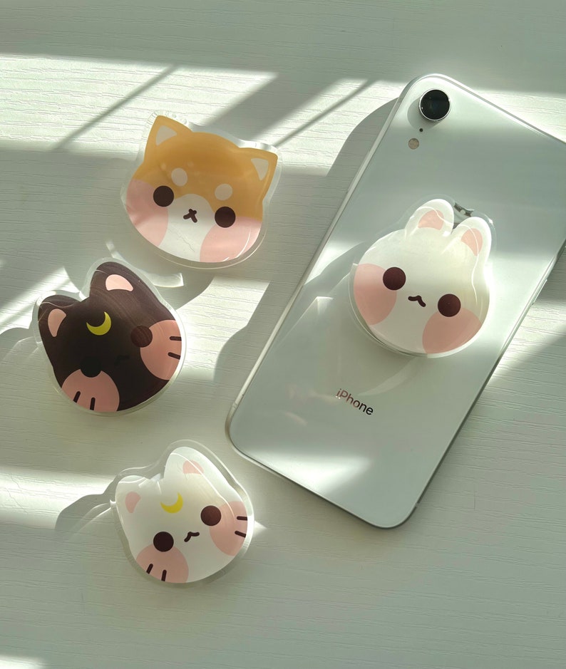 May include: Four phone grips with cute animal faces. The grips are clear plastic with a white background and pink cheeks. The animals are a brown and white dog, a black and white cat, a white cat, and a white bunny.