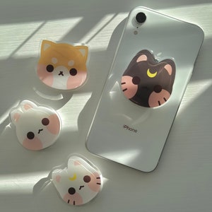 May include: Four phone grips with cute cartoon animal designs. The grips are clear with a white background and feature a brown dog, a black cat, a white cat, and a white bunny. The phone grips are on a white surface. The text "iPhone" is visible on a white phone.