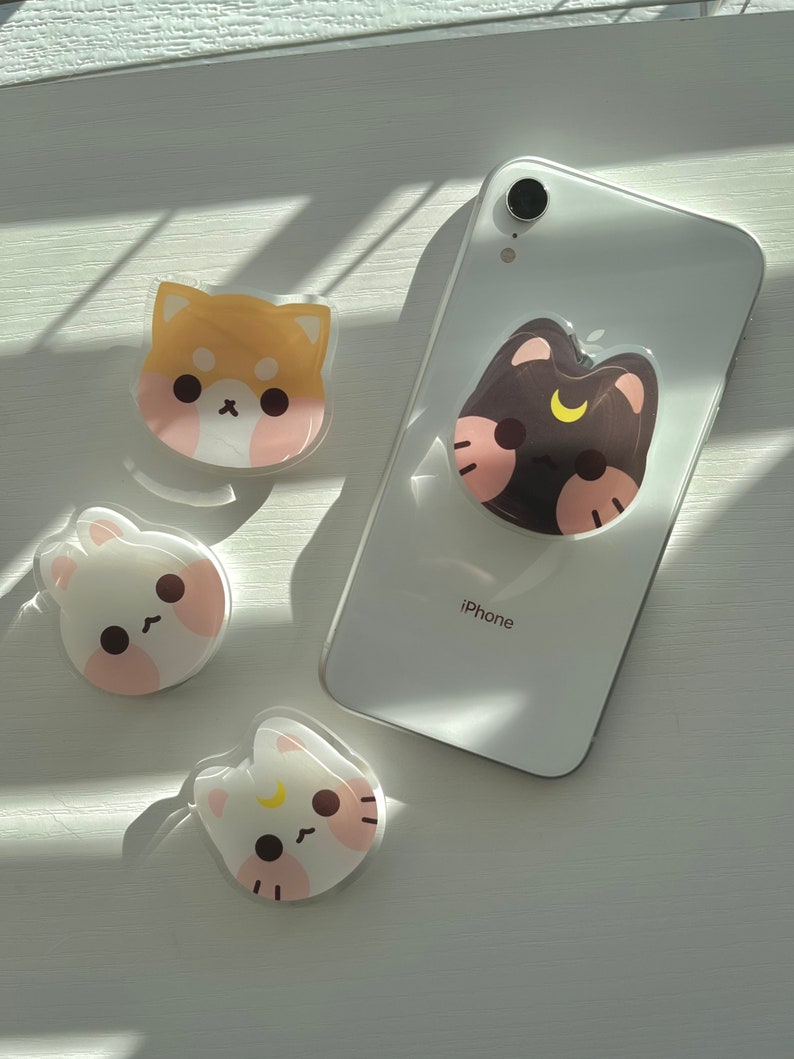 May include: Four phone grips with cute cartoon animal designs. The grips are clear with a white background and feature a brown dog, a black cat, a white cat, and a white bunny. The phone grips are on a white surface. The text "iPhone" is visible on a white phone.