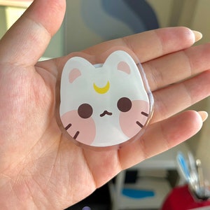 May include: A clear, round sticker featuring a white cat design. The cat has pink cheeks, brown eyes, and a yellow crescent moon on its forehead.