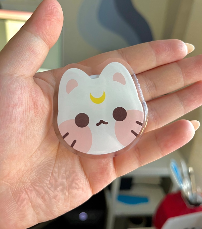 May include: A clear, round sticker featuring a white cat design. The cat has pink cheeks, brown eyes, and a yellow crescent moon on its forehead.