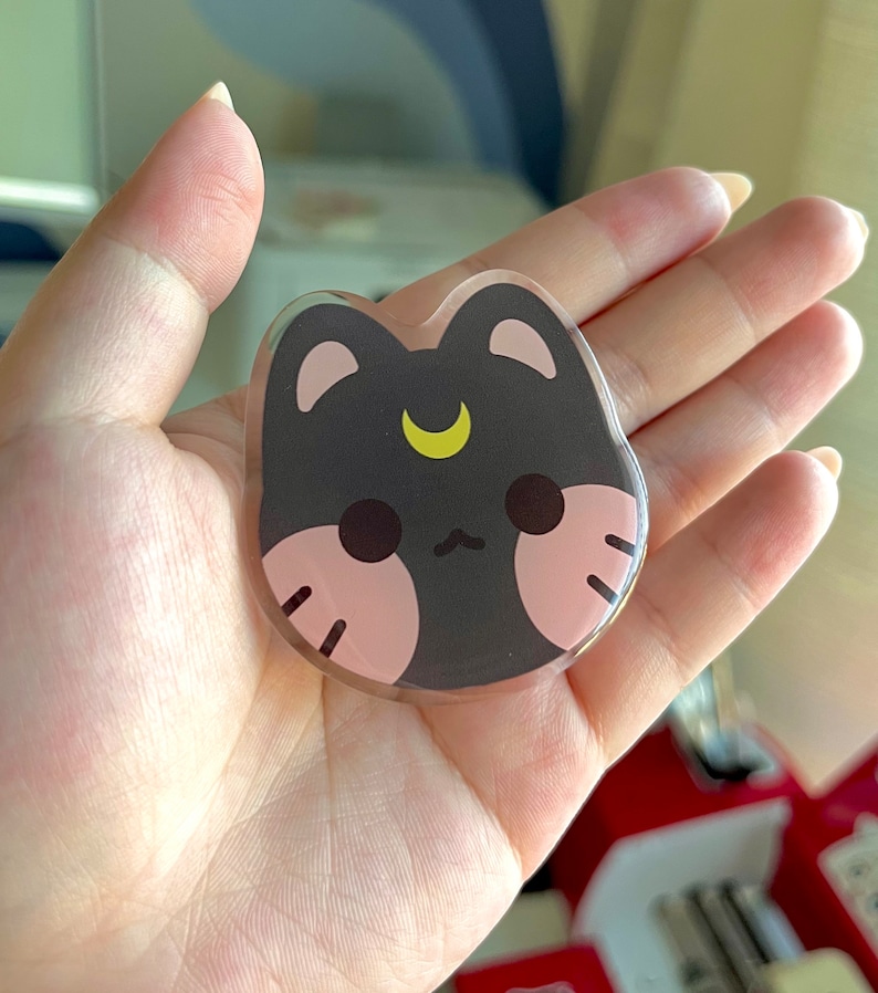 May include: A black and pink cat with a crescent moon on its forehead. The cat has a cute, cartoonish style and is made of clear acrylic.