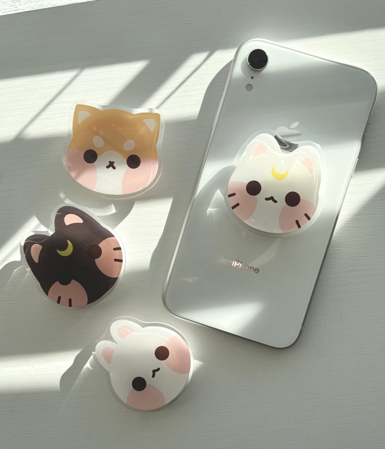 May include: Four phone grips with cute animal designs. A white cat with a crescent moon, a brown dog, a black cat with a crescent moon, and a white bunny.