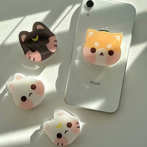 May include: Four phone grips with cute animal designs. The grips are clear with a white background and feature a black cat, a brown and white dog, a white rabbit, and a white cat. The grips are attached to a white iPhone.