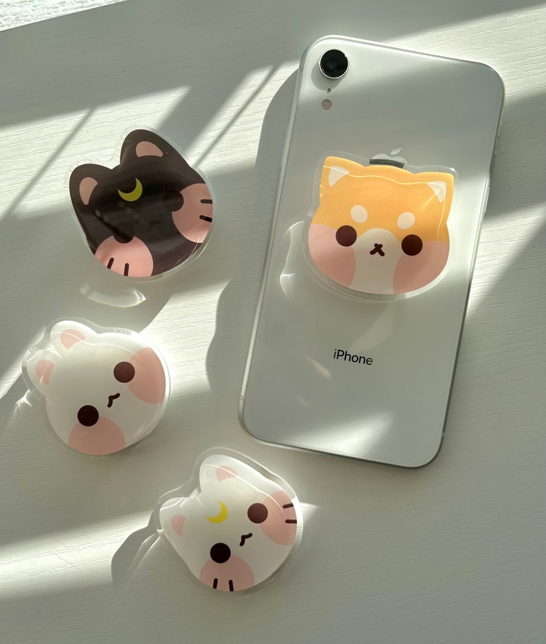 May include: Four phone grips with cute animal designs. The grips are clear with a white background and feature a black cat, a brown and white dog, a white rabbit, and a white cat. The grips are attached to a white iPhone.
