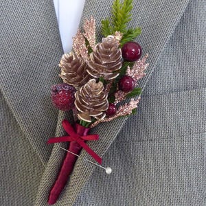 May include: A decorative buttonhole with brown pinecones, burgundy berries, and green foliage, embellished with glitter and a red ribbon. The floral arrangement is pinned to a grey suit jacket, offering a classic and elegant appearance.