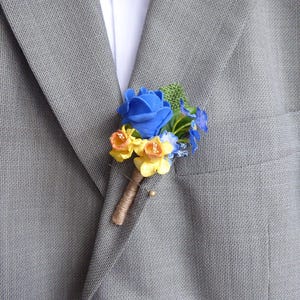 May include: A blue rose and yellow flower buttonhole pinned to a grey suit jacket. The buttonhole has a brown stem wrapped in twine and a gold pin. The suit jacket has a pocket on the left side.
