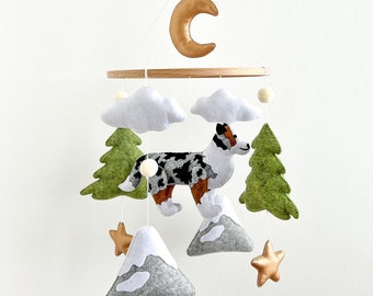 Könnte beinhalten: Ein Filz-Babymobil mit einer Sichelmond, Wolken, einem Hund, Bäumen, Bergen und Sternen. Der Hund ist ein Blue Merle Australian Shepherd. Das Mobile ist im Wald-Thema gehalten.