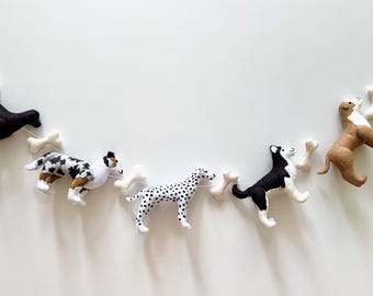 May include: A garland of felt dogs with white bone accents. The dogs are black, brown, white, and black and white spotted. The garland is strung on white twine.