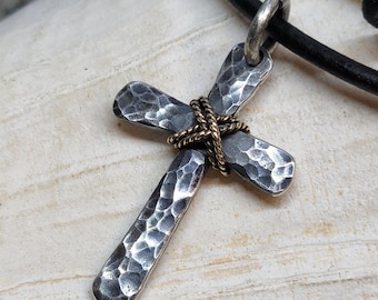 May include: A silver hammered cross pendant with a gold wire wrap, hanging from a black leather cord.