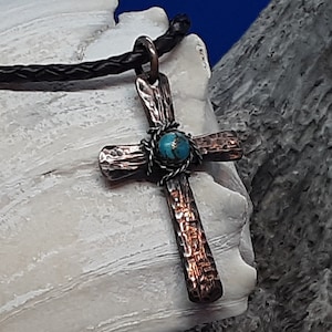 May include: A hammered copper cross pendant with a turquoise stone inlay, hanging on a black leather cord.