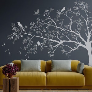 May include: A gray silhouette of a tree with branches and leaves, with small birds perched on the branches. The tree is on a dark gray wall behind a yellow couch with gray throw pillows.