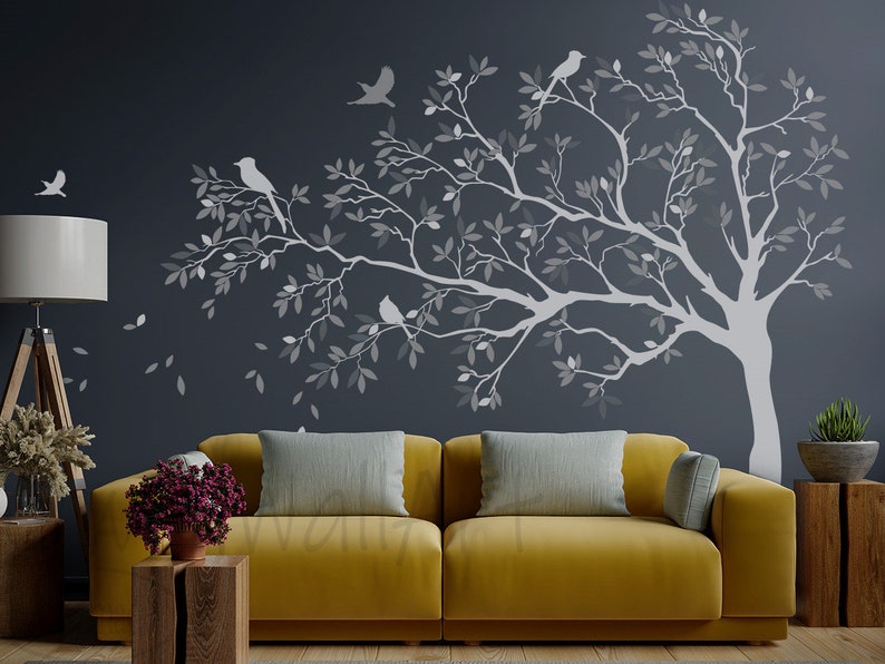 May include: A gray silhouette of a tree with branches and leaves, with small birds perched on the branches. The tree is on a dark gray wall behind a yellow couch with gray throw pillows.