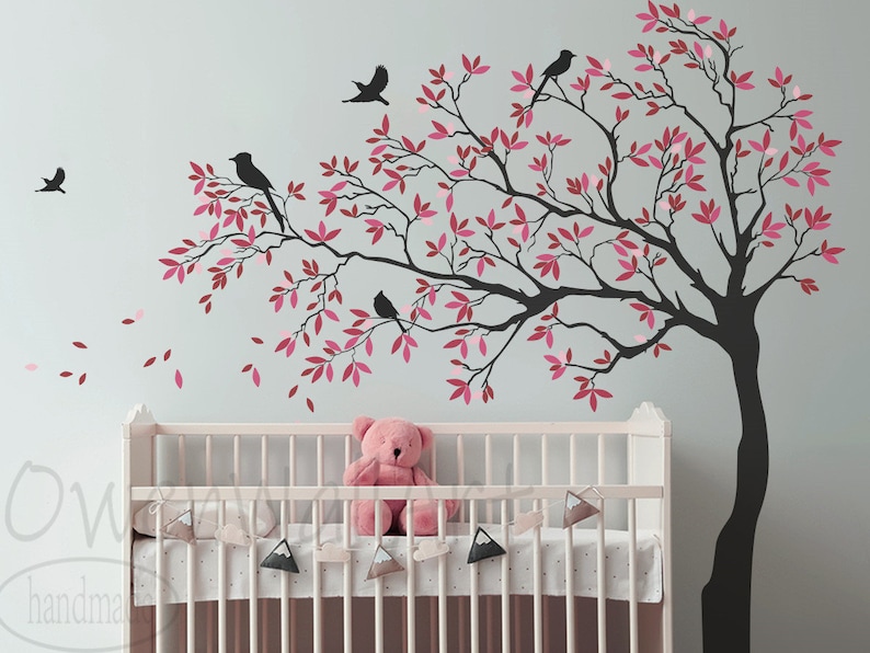 May include: A black silhouette of a tree with pink leaves and birds on the branches. The tree is on a light blue wall above a white crib with a pink teddy bear inside.