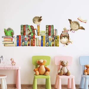 Op de afbeelding: Een witte muur met een boekenkaststicker met kleurrijke boeken en bruine uilen die boeken vasthouden. Op de voorgrond staan drie kinderstoelen, elk met een teddybeer erop. De stoelen zijn groen, roze en blauw.