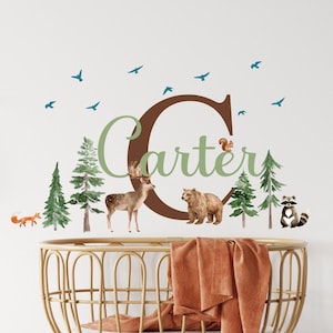 Könnte beinhalten: Wandtattoo für das Kinderzimmer mit einem braunen Buchstaben "C" und dem Namen "Carter" in grüner Schrift. Das Design umfasst Aquarellillustrationen von Waldtieren wie Fuchs, Hirsch, Bär und Waschbär sowie grüne Bäume und blaue Vögel.