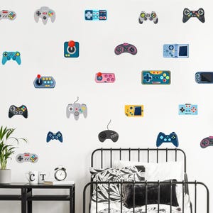Puede incluir: Una pared blanca decorada con varias pegatinas coloridas de mandos de videojuegos. Los mandos tienen diferentes formas y colores, incluyendo azul, gris, rosa y amarillo. Se ve una estructura de cama y una mesita de noche de metal negro.