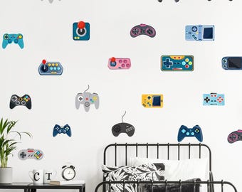 Op de afbeelding: Een witte muur versierd met diverse kleurrijke stickers van gamecontrollers. De controllers hebben verschillende vormen en kleuren, waaronder blauw, grijs, roze en geel. Een zwart metalen bedframe en nachtkastje zijn zichtbaar.