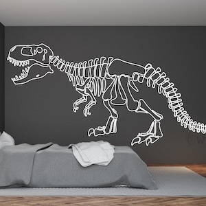 May include: A white dinosaur skeleton decal on a dark gray wall. The dinosaur is facing right and has its mouth open. The decal is in a bedroom setting with a gray bed and a wooden floor.